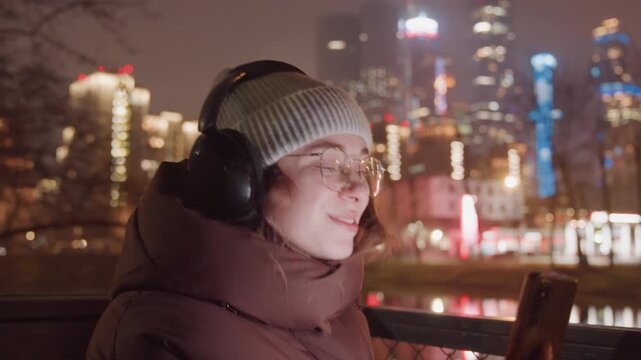 Recording young woman with smartphone city, riverfront backdrop, capturing vlog footage, headphones on, glasses reflecting neon, animated gestures, editready clips, creator energy
