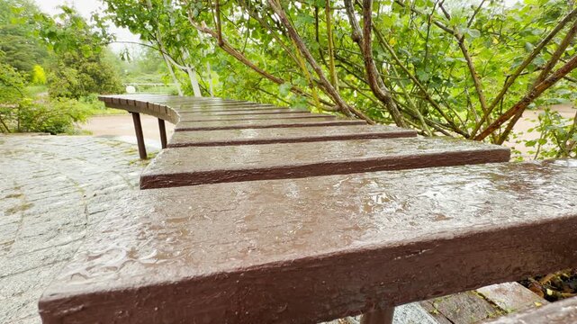 A curved wooden bench sits empty as soft rain falls in a green park. Shimmering water puddles and droplets coat the brown wooden slats next to wet stone pathways.