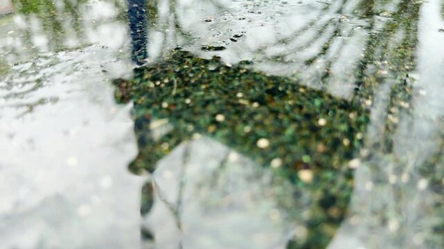 A dark rain puddle reflects a swinging swing set frame on a textured playground floor. The water surface shimmers gently during a quiet, gloomy afternoon outdoors.