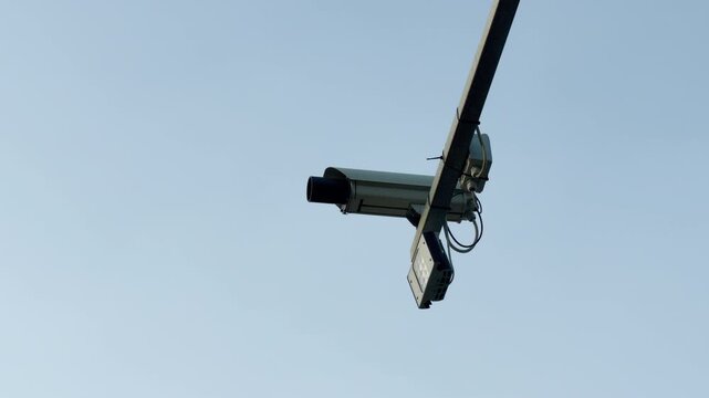A traffic enforcement camera is attached to a high metal beam. The system points downward to watch the road below. It operates under a bright, cloudless sky.