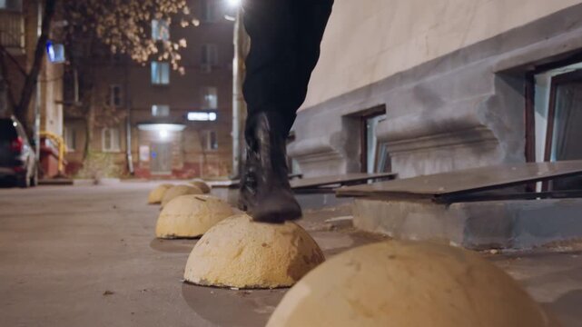 Urban alley walker hopping over bollards, narrow street scene with worn yellow domes, confident black boots stepping in rhythm, casual bag swing, lamplight reflections on asphalt, quick sequence