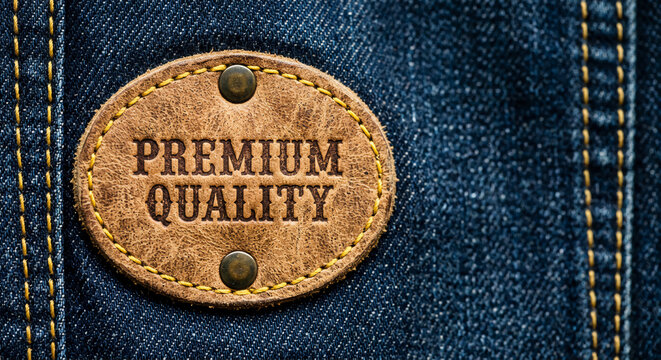 An oval, textured brown leather patch stamped with "PREMIUM QUALITY" is riveted onto dark blue denim fabric with yellow stitching.
