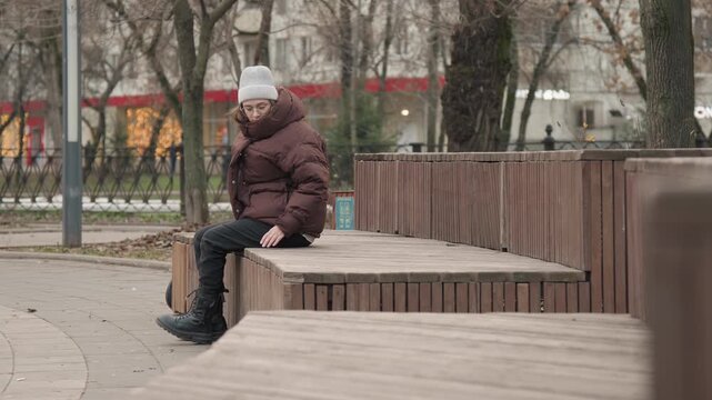 Caucasian woman waiting alone bench in winter park, layered coat and knit hat, sitting with phone and boots, reflective posture and quiet urban scene, soft ambient lights and stillness