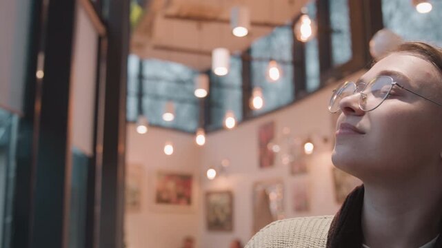 White woman reflecting inside gallery with warm bulbs and modern paintings, glasses catching ceiling lights, contemplative student or artist studying exhibits, quiet creative atmosphere and soft bokeh