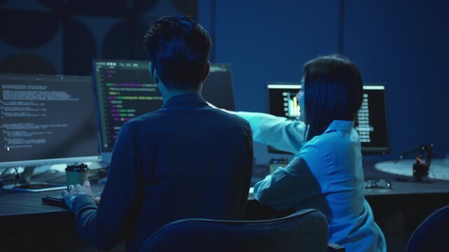 Two young software developers work side by side writing code on computer monitors in a dark blue lit modern office at night, focused on a software programming project.