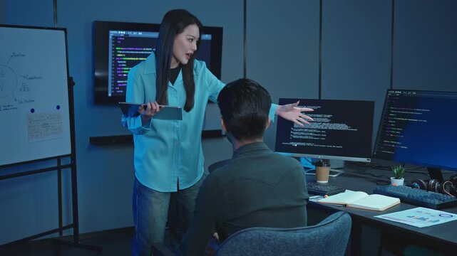 Young female software developer presents ideas at a whiteboard while explaining code to a seated male colleague in a dark blue lit modern technology office at night.
