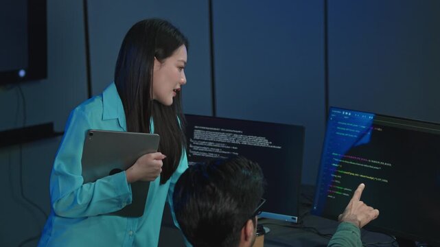 Smiling young female developer holds a tablet while collaborating with a male coworker at a desk full of code monitors in a dark blue lit modern technology office.