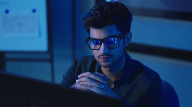 Young male software developer wearing glasses focuses intently on his computer screen while coding late at night in a dark blue lit modern technology startup office.
