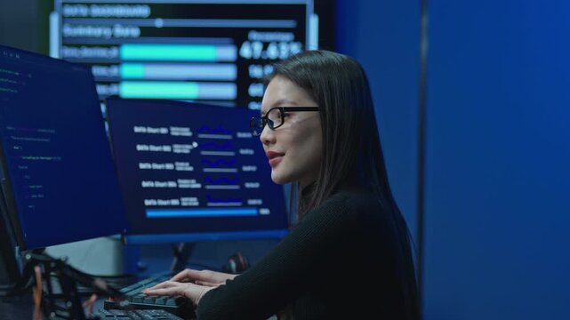 Young female developer works focused at a desk with multiple code and data monitors in a dark blue lit modern technology office during a late night work session.