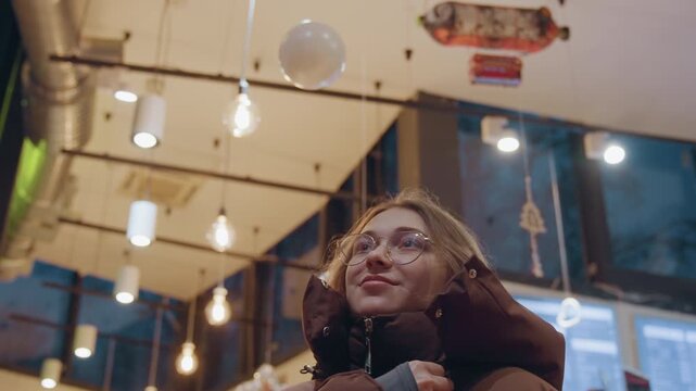 White woman looks up at lights, hooded puffer and dangling bulbs create warm overhead glow, joyful gaze and small smile, holiday gallery atmosphere, brief moment of wonder and pause, cinematic wide