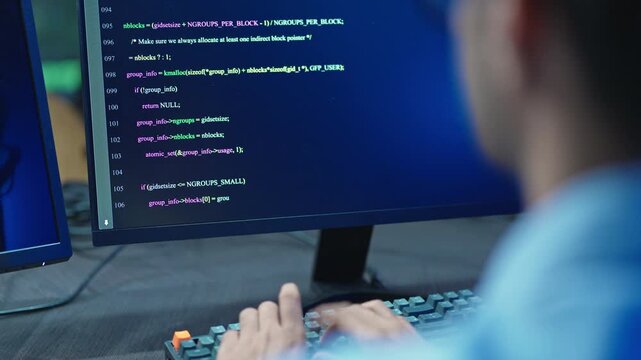Close up of a software developer working at a computer with lines of program code displayed on the monitor in a dark blue lit modern technology office at night.