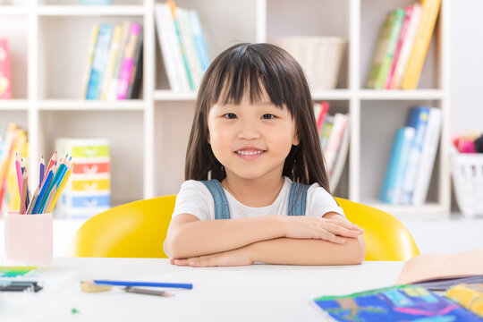 A polite and happy little Asian girl sitting up straight at her desk with crossed arms and smiling brightly at the camera.