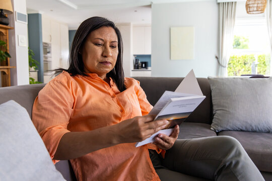 Mature adult Asian woman sitting on gray sofa at home reading folded booklet in orange shirt