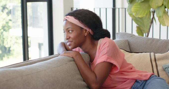 African woman sitting in pink top on sofa by tall windows leaning shifting smiling engaging camera