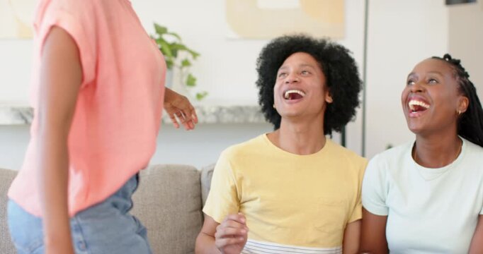 Friends laughing on gray sofa as friend coming from left greeting and hugging, bracelets visible