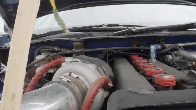 Wideangle turbo engine bay showing blue hood reflection and large intake piping red coilpacks and intercooler lines visible, track tuner prepping car for street event, open bonnet perspective, outdoor