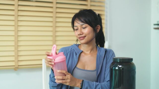 Young Asian woman in a blue jacket smiles while holding a pink protein shaker bottle in a modern kitchen, preparing a nutritious supplement drink at home in soft natural light.