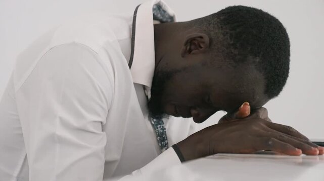 Defeated man with head bowed. Black man sits postdate in private remorse, hands folded on table, heavy breathing and introspective silence white shirt and tie, intimate closeup of exhaustion, grief
