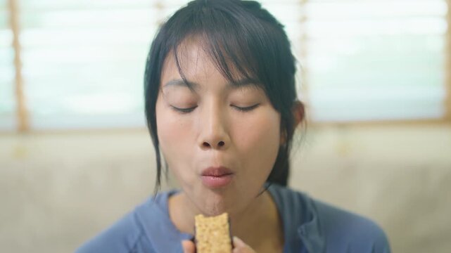 Young Asian woman happily chews a healthy granola energy bar with her eyes closed, enjoying a tasty wholesome snack in a bright comfortable living room in soft natural light.