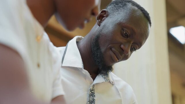 Black couple reading paper together, tutor guiding student on veranda, warm natural light, white shirts and tie, close intimate conversation and thoughtful smiles, gentle guidance and supportive