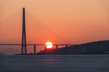 Obraz premium Sunset view behind a suspension bridge with cables silhouetted against an orange sky over a calm body of water reflecting the warm hues of dusk
