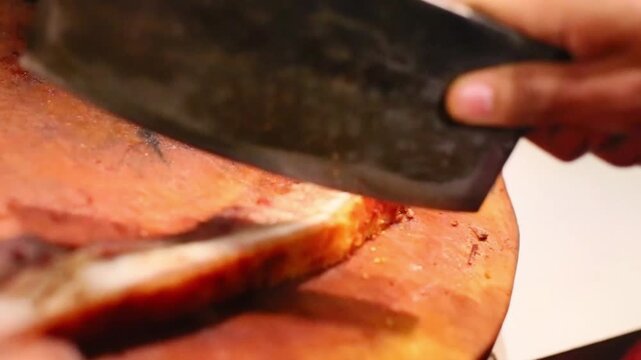 Close-up image of a butcher knife slicing crispy roast pork on a wooden cutting board in a food preparation scene in a kitchen or street food stall.