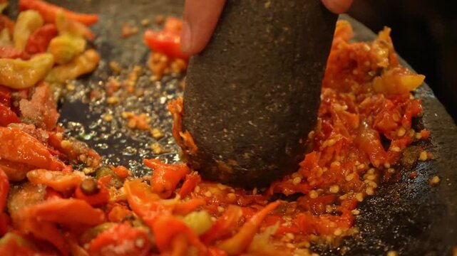 Fresh red chili sambal being crushed on a stone mortar, spicy Indonesian sauce close-up with bold color and rustic texture.