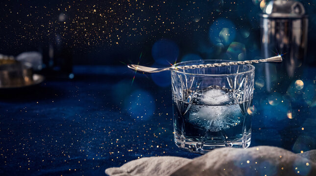 Elegant cocktail glass with a silver spoon and ice, reflecting sparkling lights on a deep blue background.