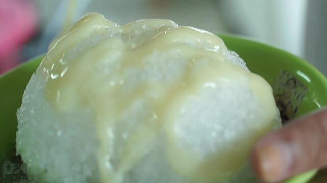 Sweet condensed milk poured over shaved ice in a green bowl, refreshing Asian dessert close-up with creamy texture.