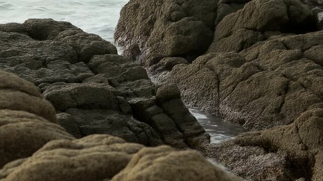 Static slow motion pan tracking of rocky shoreline with waves lapping into tide pools on overcast coastal daytime