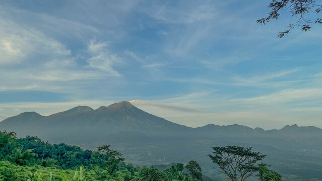 Beautiful panoramic view of Mount Arjuno and Mount Welirang in East Java, Indonesia. The landscape features lush tropical vegetation, volcanic mountain peaks, soft atmospheric haze, and dramatic cloud
