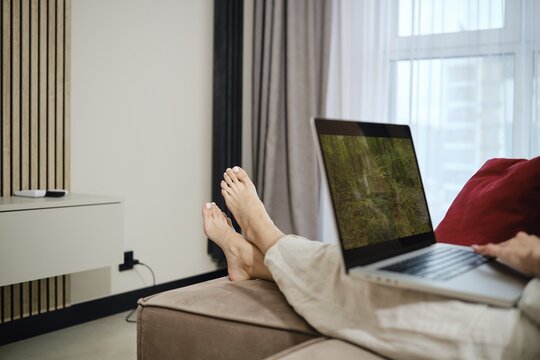 In a modern living room, a barefoot woman sits comfortably on a couch, using a laptop while lounging