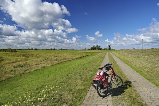 A bicycle stands on a paved path leading through a vast meadow landscape under a cloudy sky, dyke, cycle tours, cycle tourism, Gartow, Lower Saxony, Germany