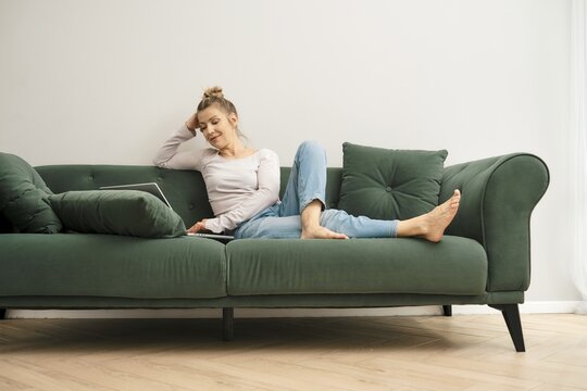 In a cozy living room with natural light, a middle-aged woman sits comfortably on a green couch and chatting on her laptop