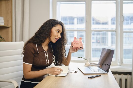 A young woman is actively budgeting at her desk, holding a pink piggy bank in one hand while writing notes in a notebook with her other hand