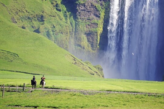 Water masses plummeting vertically into the depths, reflection in the water, green landscape, two riders on Icelandic horses, Skogafoss, Iceland
