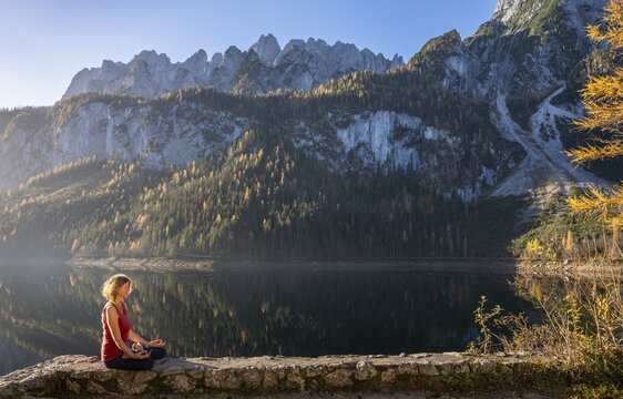 A woman doing yoga by the lake. Lotus position (Padmasana) . Vorderer Gosausee in autumn with a view of the Dachstein mountains. The Gosaukamm on the right. Blue sky, good weather. Reflection. Morning atmosphere. Vorderer Gosausee, Gosau, Gosau valley, Salzkammergut, Upper Austria, Austria