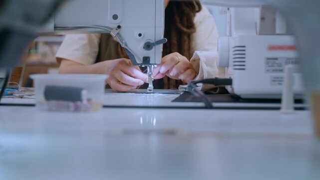 A young woman works in a sewing studio, stitching fabric with an industrial sewing machine. She focuses intently on the seam allowance, keeping the fabric aligned with the guide lines