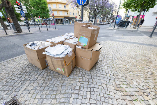 Piles of cardboard boxes filled with discarded paper waste on a cobblestone street corner in an urban area surrounded by trees and buildings