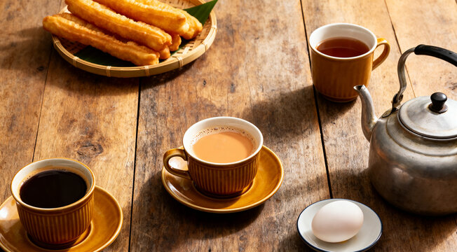 Traditional Thai hot tea with milk on the wood table there are Traditional Thai hot coffee, deep-fried doughstick (patongko), hot tea, Kettle and soft-boiled egg plated around