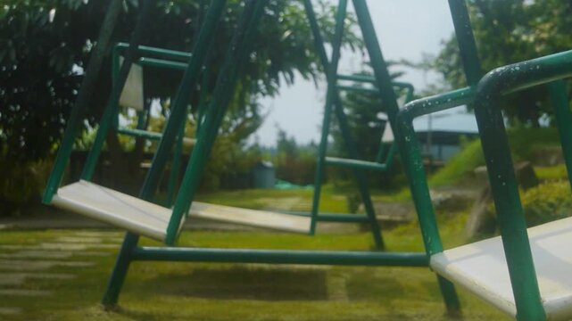 Empty swings sit quietly within a green metal playground frame, hinting at leisure and childhood memories amidst lush outdoor surroundings
