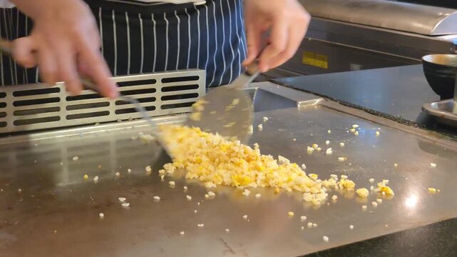 Eye Level Close Up View of Professional Teppanyaki Chef Using Two Spatulas to Stir-Fry Rice on a Hot Flat Griddle