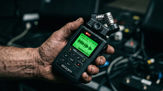 A weathered hand holds a professional black digital audio recorder featuring a bright green display screen and metallic microphone capsules during a busy recording session today.