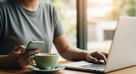 Fototapeta premium Woman sitting near window with coffee cup and smartphone while typing on laptop computer working remotely in cozy cafe environment with blurred natural background