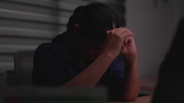 Despair in the Dim Light: A person sits with their head bowed in distress, hands clasped together, in a dimly lit room, conveying a profound sense of sadness and struggle.