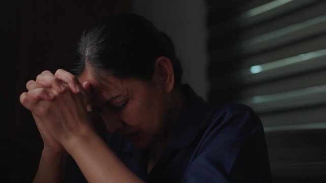 Deep Contemplation: A woman clasps her hands in prayer, her brow furrowed in deep thought, silhouetted against soft light, conveying a moment of quiet reflection or earnest plea.