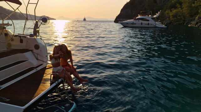 Two young women relaxing on a yacht platform at sunset, enjoying the sea, creating content and spending time together. Summer vacation, friendship, luxury boating and travel lifestyle.