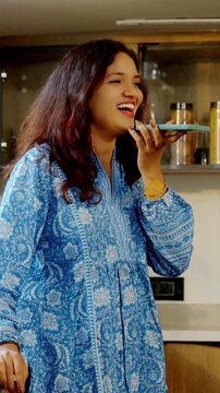 Young beautiful Indian Asian woman cooking food in a pan in the kitchen while speaking on a speakerphone, multitasking and managing kitchen tasks efficiently