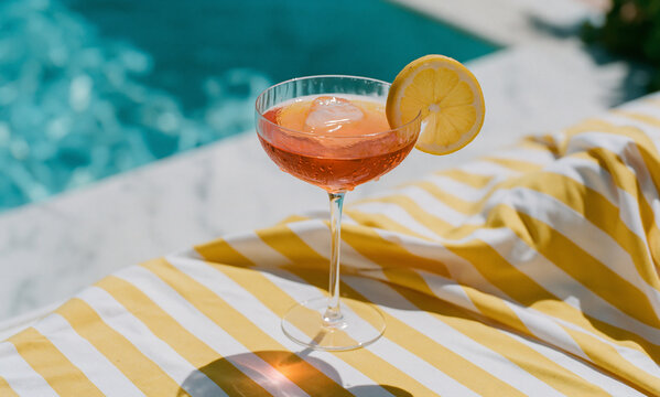 a tall coupe glass of aperol spritz with condensation droplets on a yellow and white striped daybed surface, soft focus background of turquoise pool water and white marble