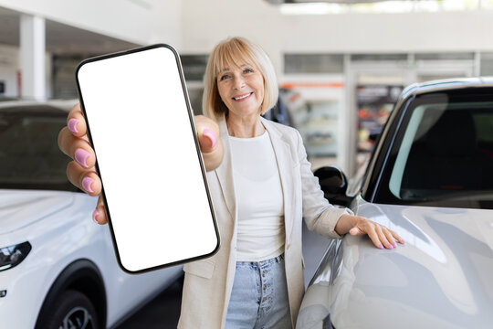 Senior woman presenting blank smartphone mockup screen while standing beside new automobile in modern dealership showroom, happy elderly female showing empty cellphone